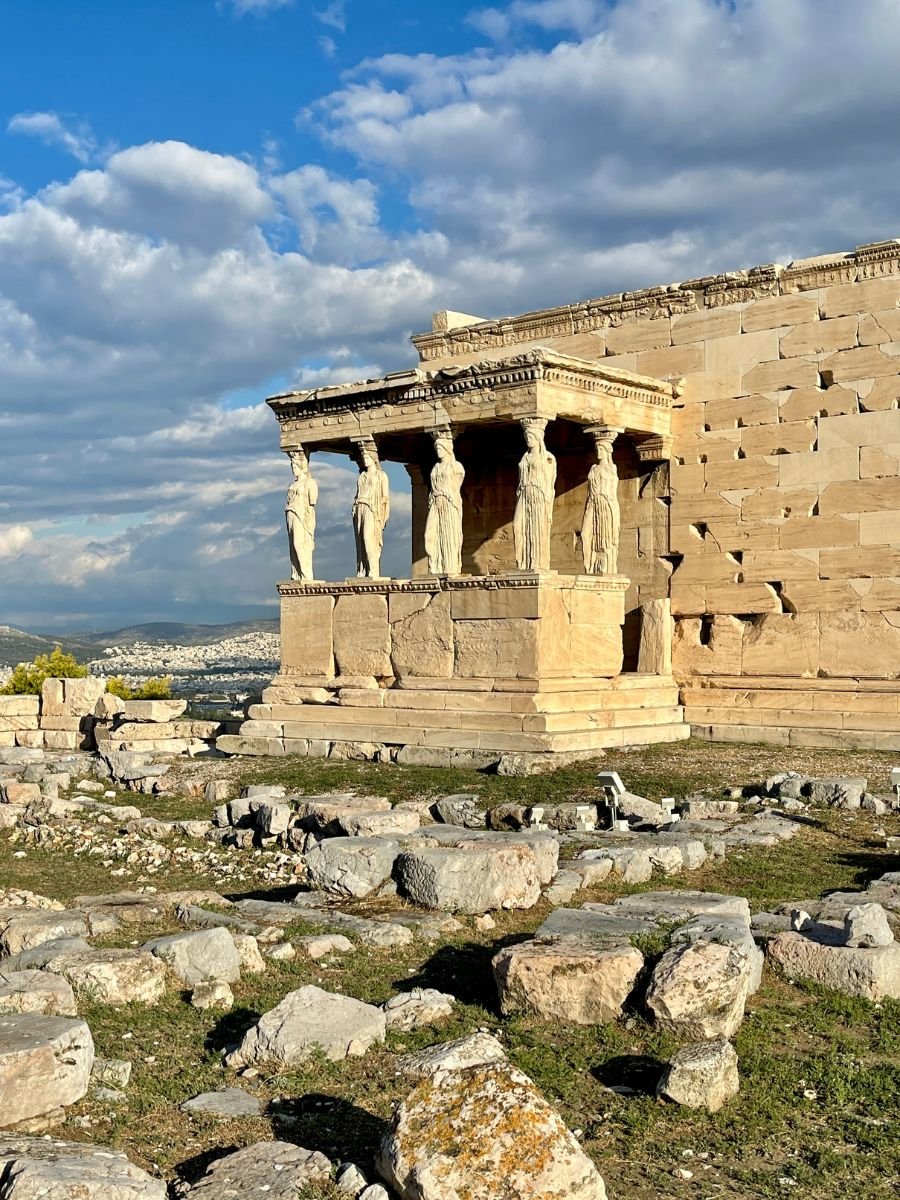Close-up of the Erechtheion temple on the Acropolis in Athens, featuring the famous Caryatids — six sculpted female figures serving as architectural columns, under a bright blue sky with scattered clouds.