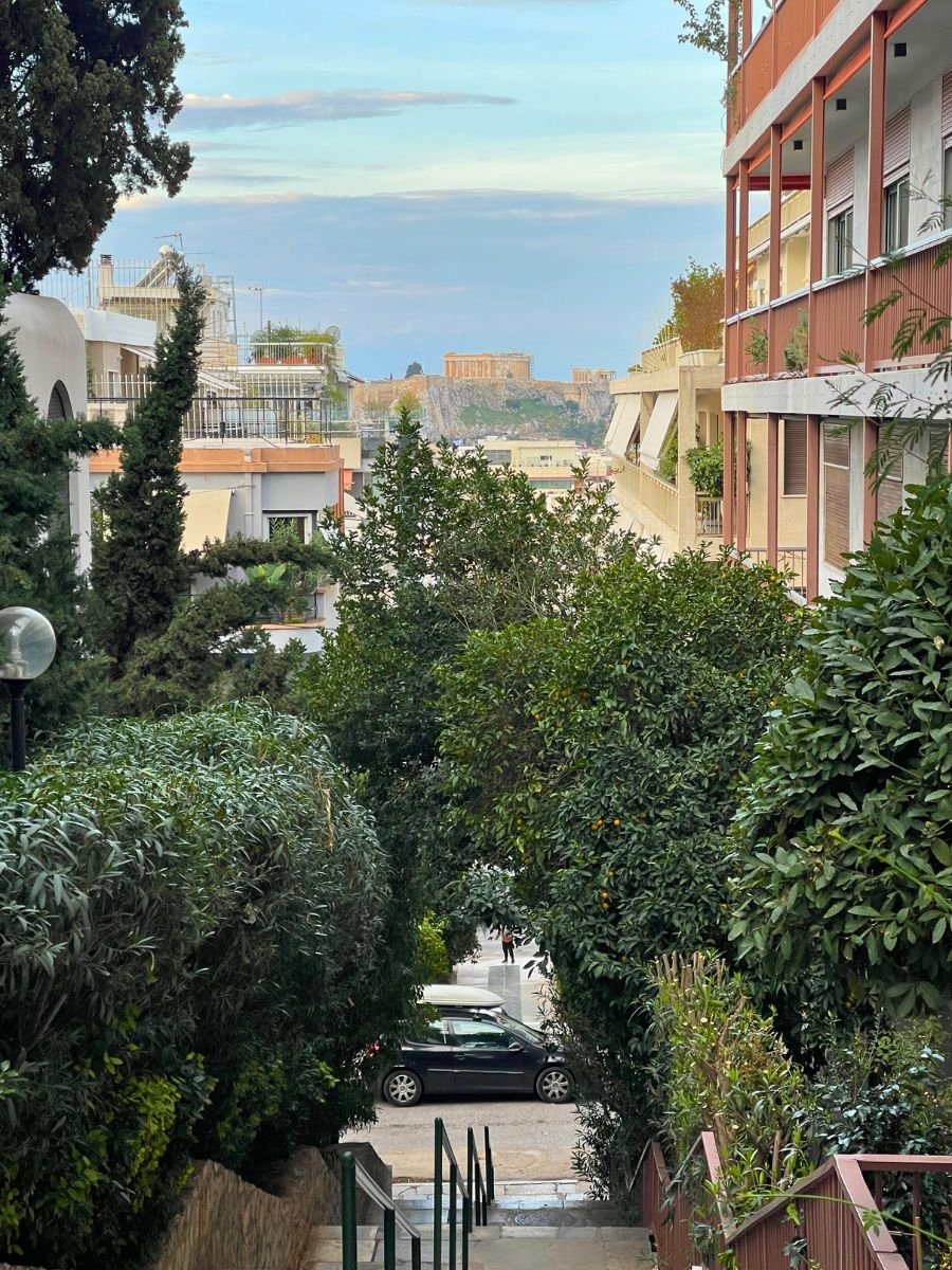 City street in Athens framed by residential buildings and lush greenery, with a distant view of the Acropolis perched on a hill under a pastel blue sky.