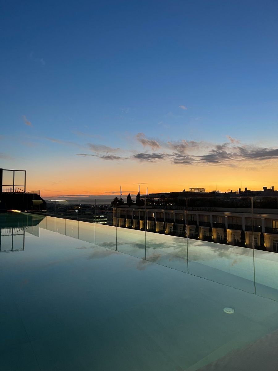 Sunset view from a rooftop infinity pool in Athens, reflecting the colorful sky and silhouetted cityscape in the water.
