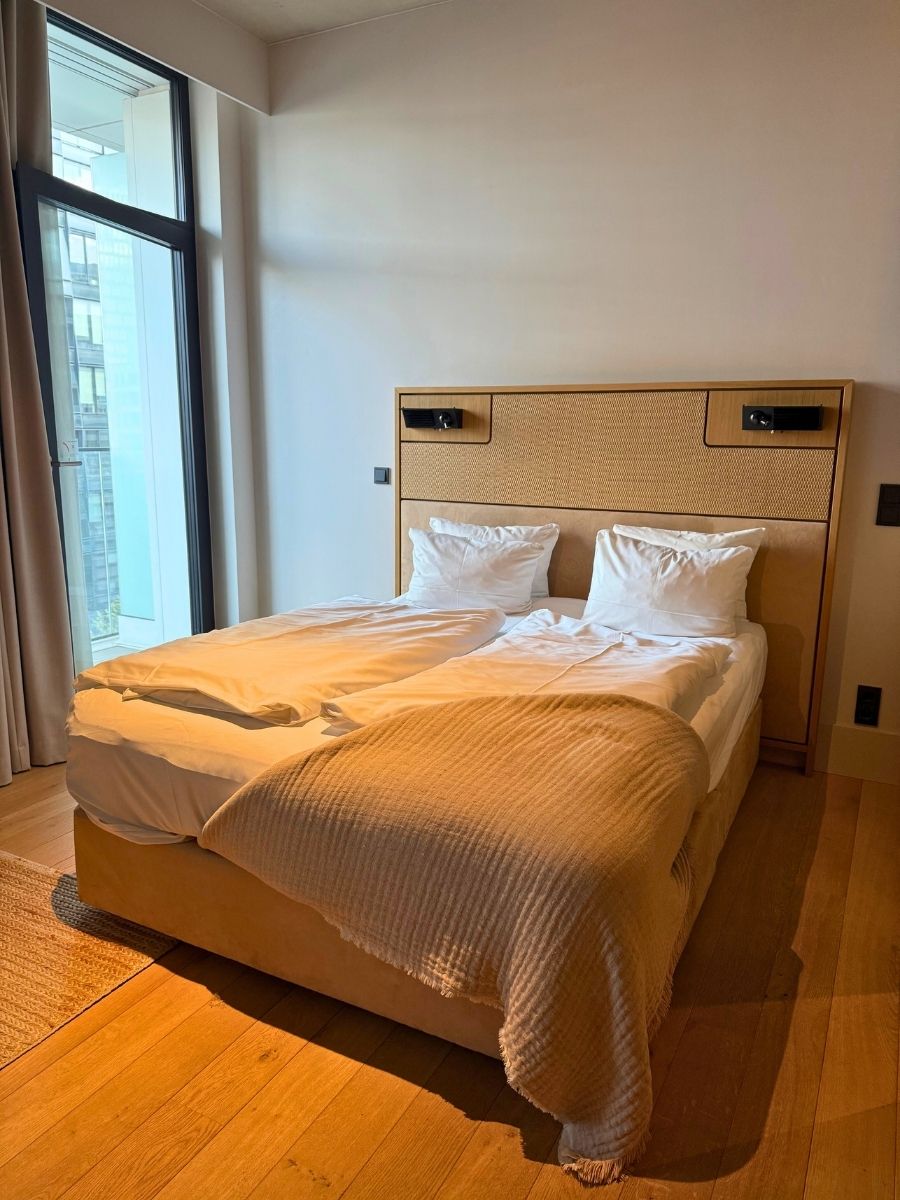 A brightly lit modern hotel room in Berlin with two separate duvets on the bed, light wood floors, and floor-to-ceiling windows.