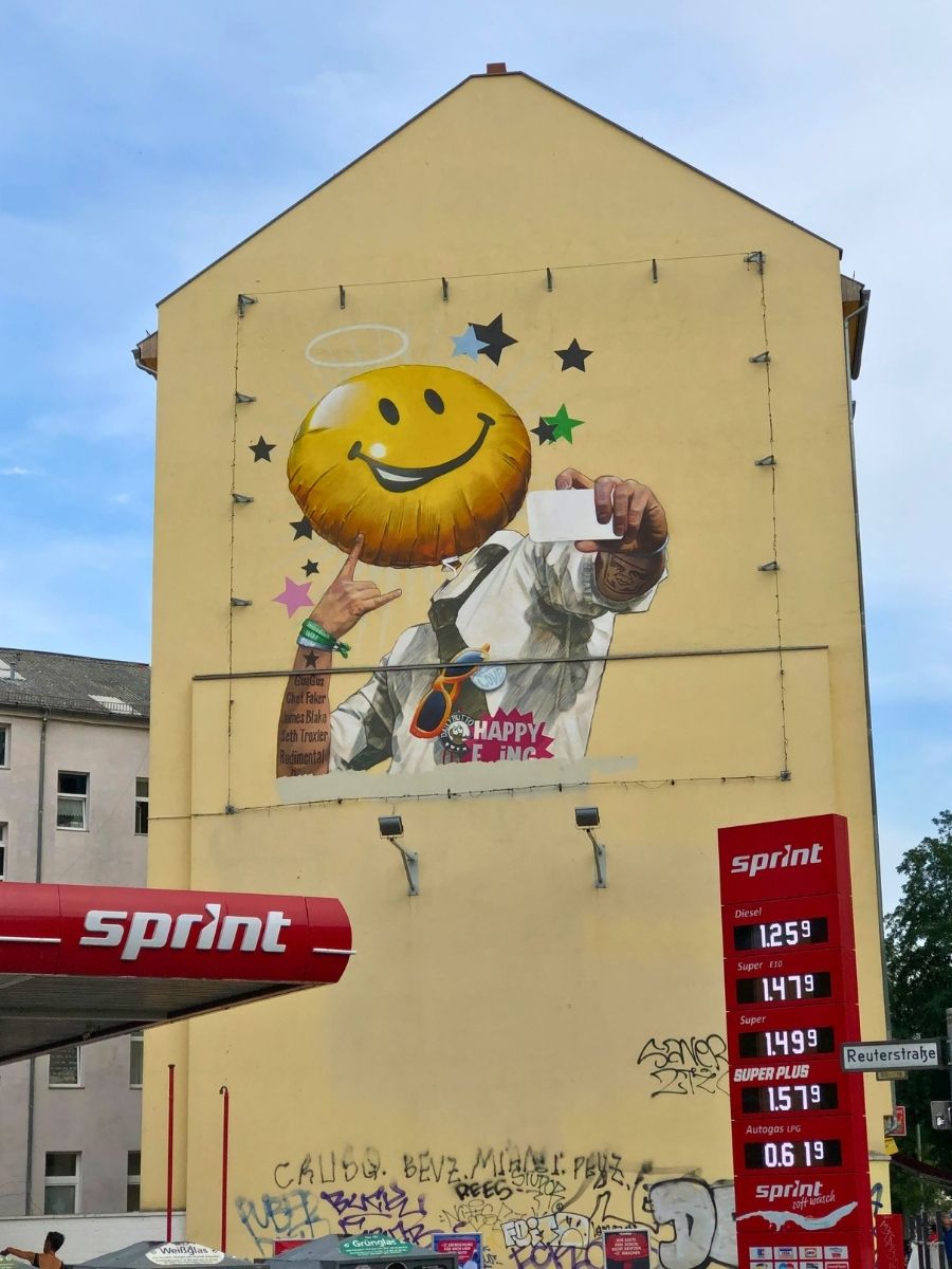 Large building mural in Berlin showing a smiley-face character taking a selfie above a Sprint gas station.