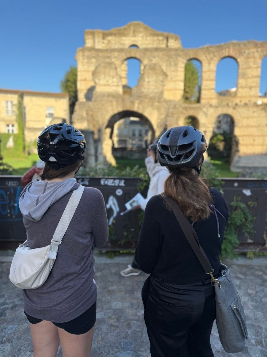 Two women in helmets facing ancient Roman ruins in Bordeaux during a guided bike tour, with graffiti-covered barriers and stone arches in view.