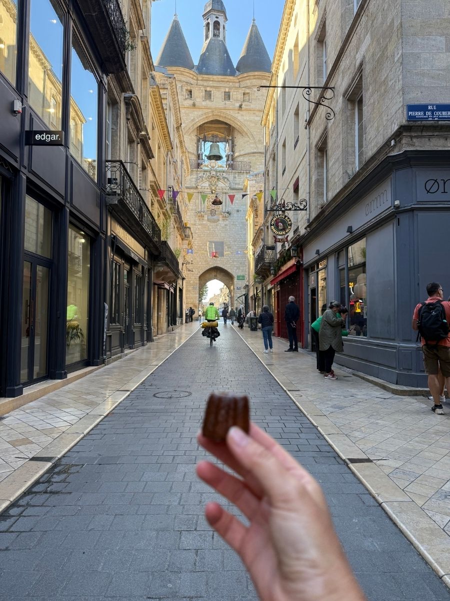 Hand holding a traditional canelé pastry with the Grosse Cloche of Bordeaux, France, and a cyclist in the background