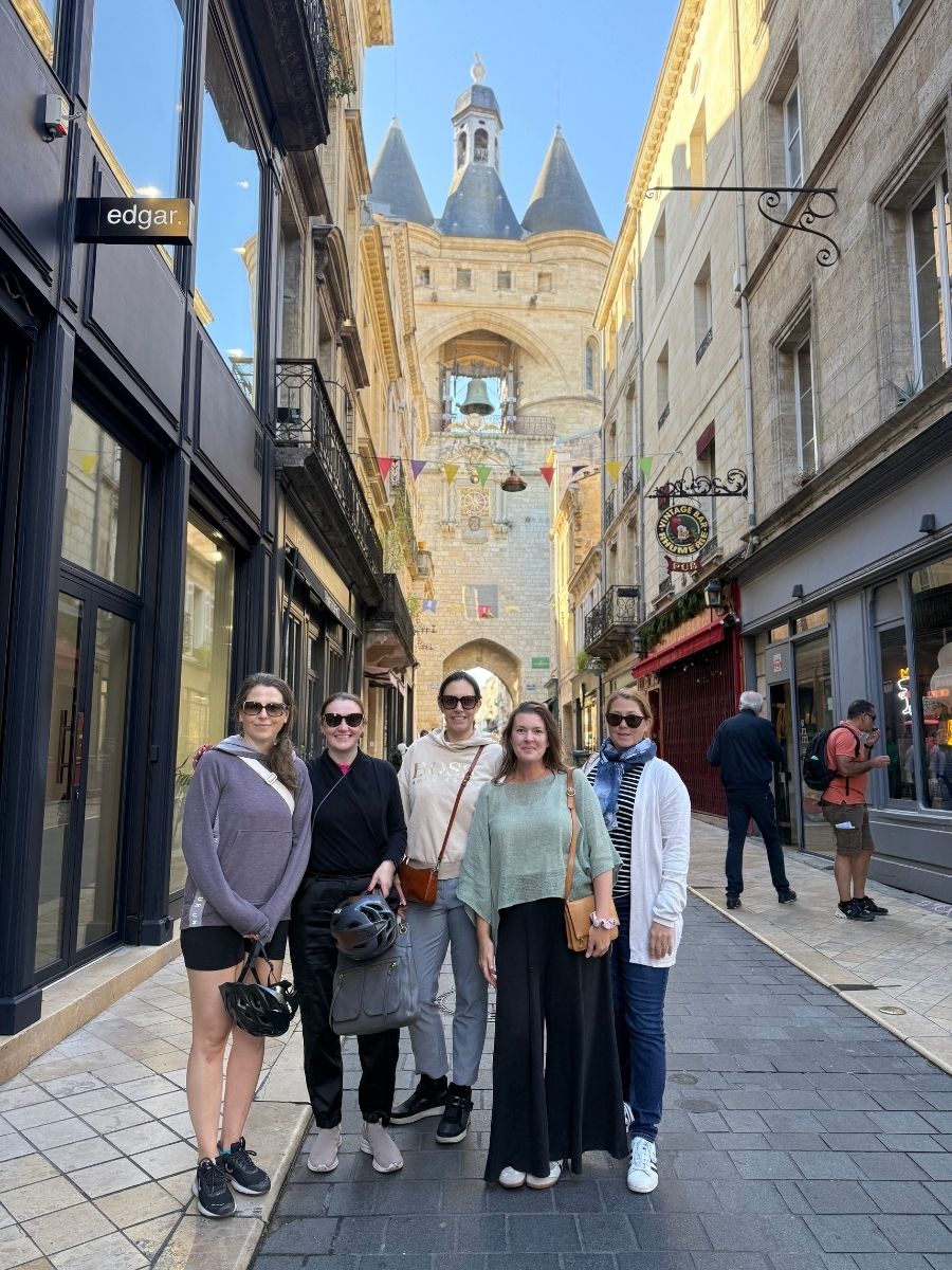 Group of five women standing on a cobblestone street in front of Bordeaux’s Grosse Cloche, a famous medieval bell tower in the heart of the city’s old town.