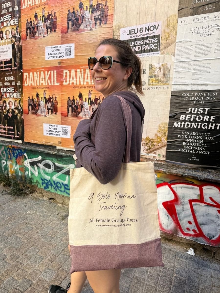 Woman smiling and walking in Bordeaux, France, carrying a tote bag that says “A Solo Woman Traveling” in front of colorful concert posters on a graffiti wall.