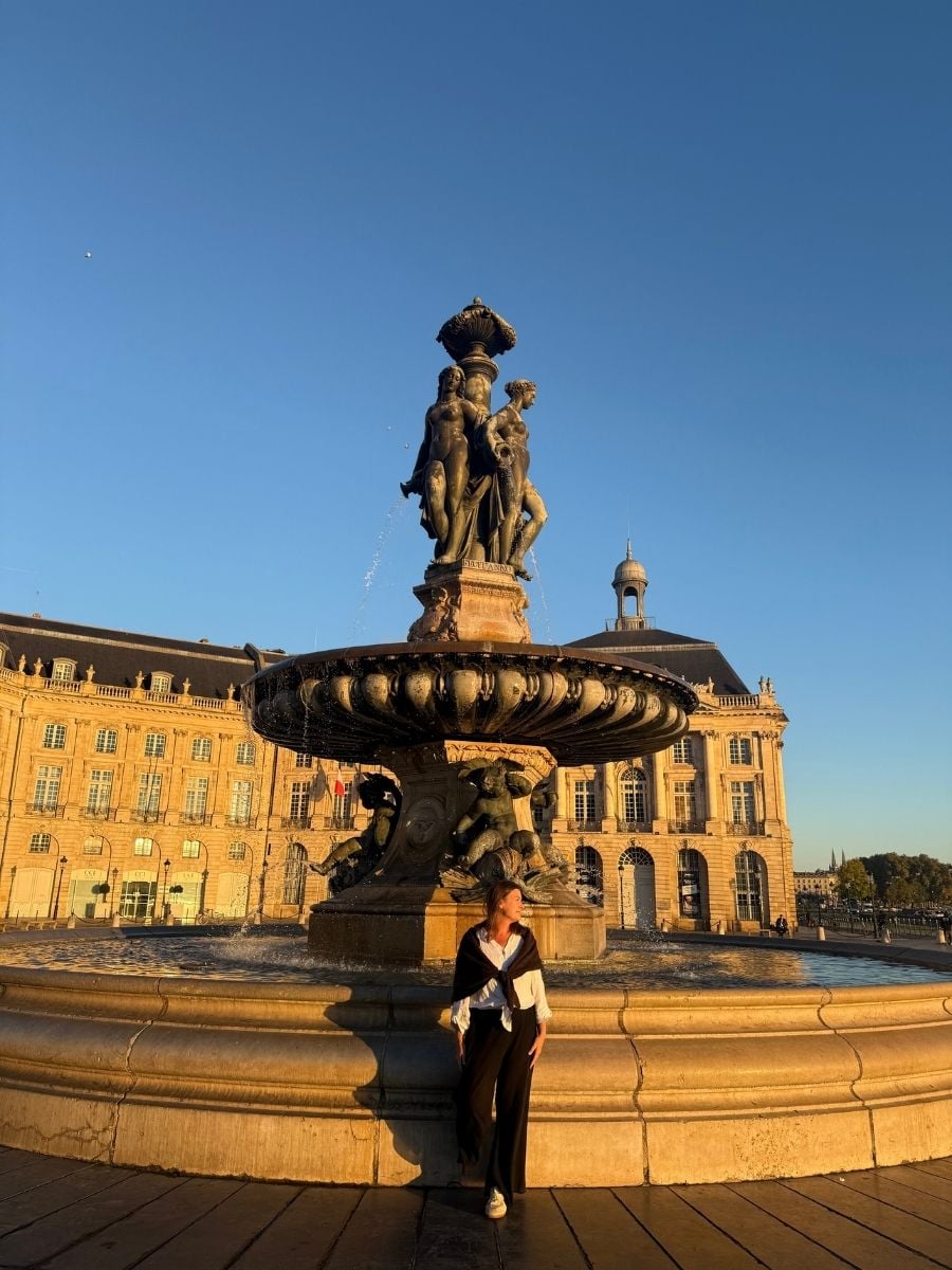 Melissa standing beside the Fountain of the Three Graces in Place de la Bourse, Bordeaux, with golden hour sunlight hitting the historic buildings.