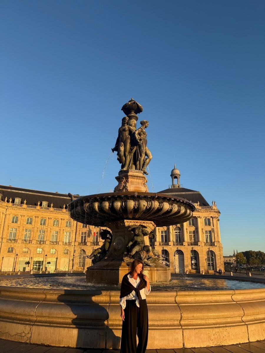 Melissa posing in front of the grand Fountain of the Three Graces in Bordeaux’s Place de la Bourse with warm sunlight casting dramatic shadows.