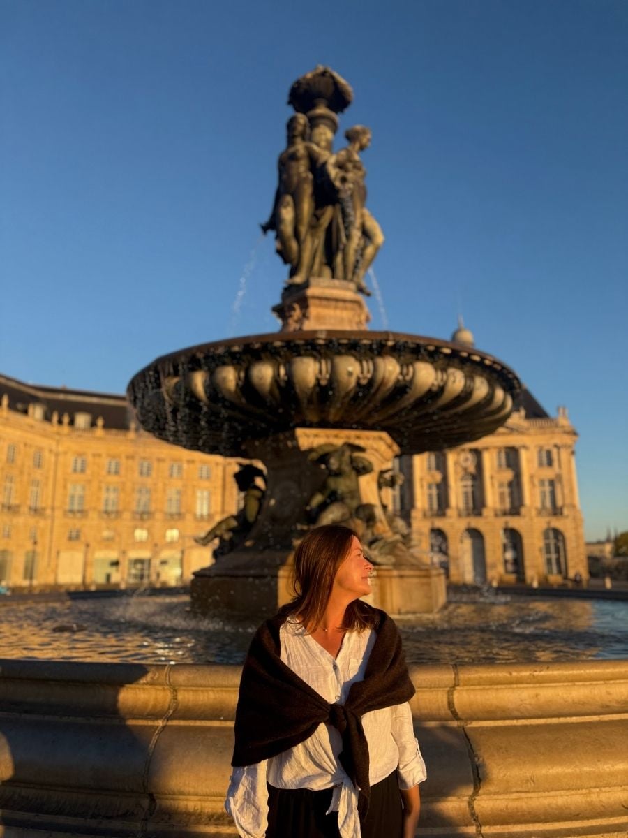 Melissa smiling while standing in front of the Fountain of the Three Graces at Place de la Bourse in Bordeaux, France during sunset.