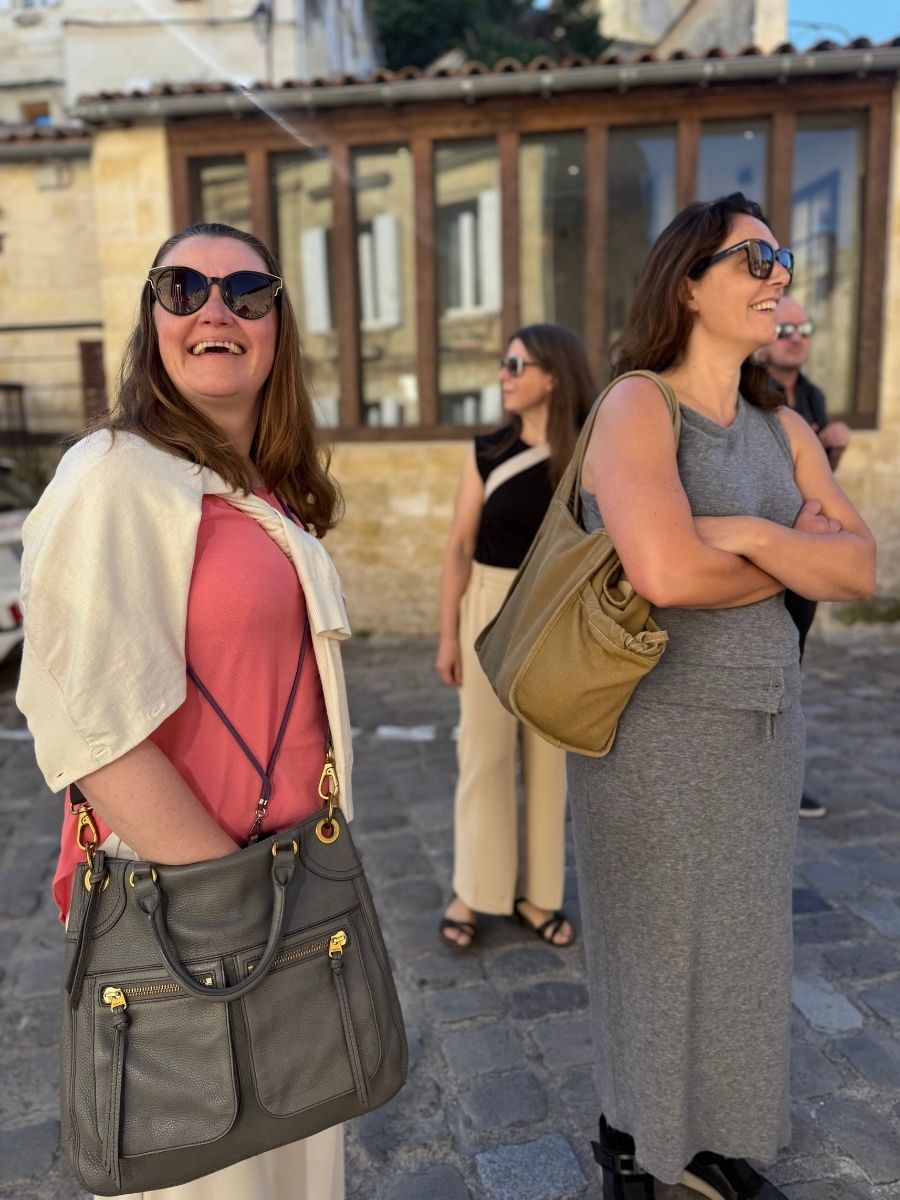 Three women smiling and chatting on a cobbled street in Saint-Émilion, Bordeaux, France, with historic limestone buildings in the background.