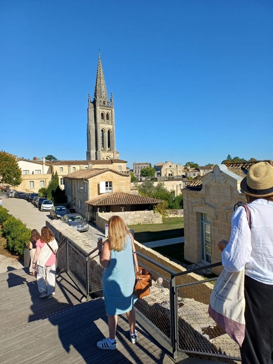 A group of women walks toward the towering bell tower of a historic church under a bright blue sky in Saint-Émilion, Bordeaux.