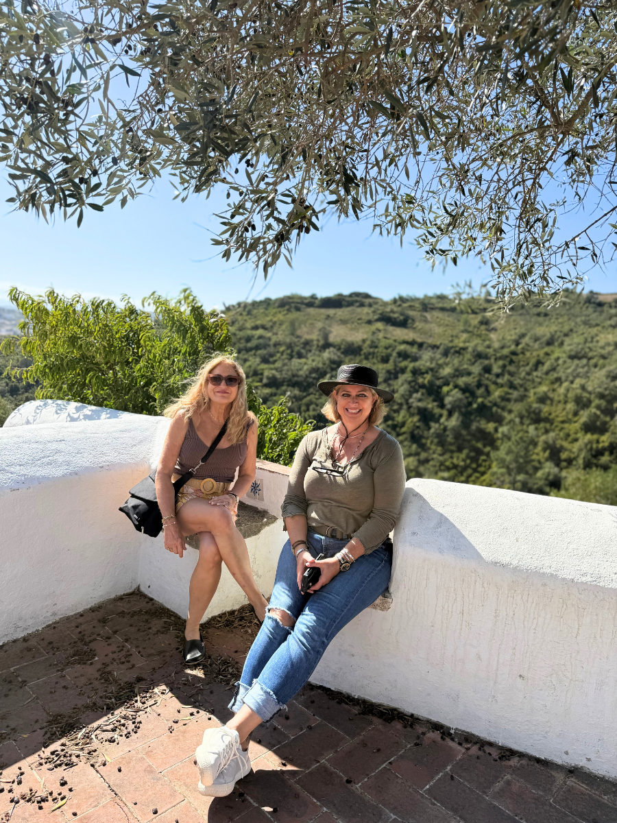 Two women smiling and posing on a low, white wall overlooking a bright, sun-drenched green hillside, likely at a vineyard or rural viewpoint in Portugal.