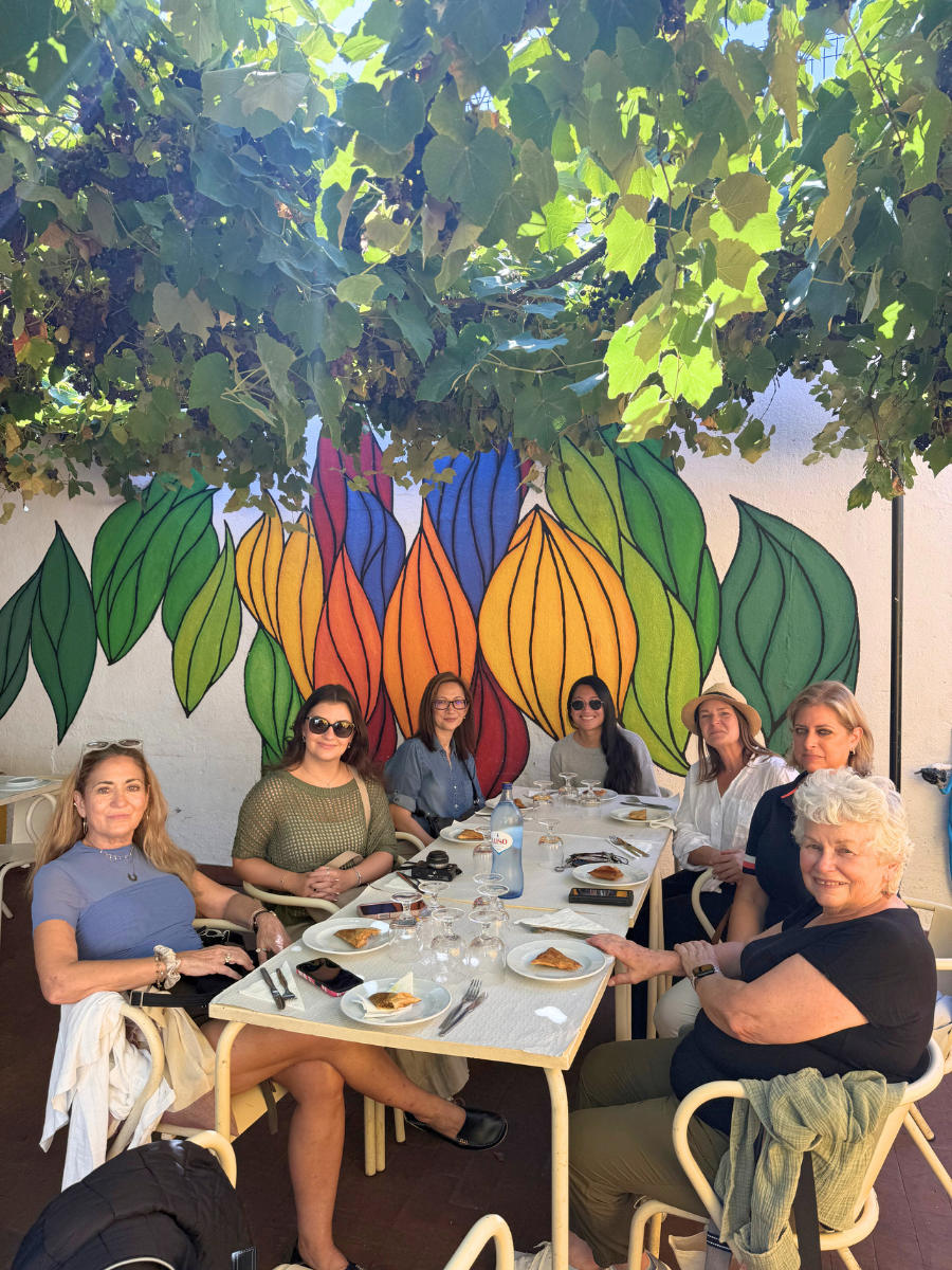 Group of seven women seated at an outdoor cafe under a lush grapevine canopy, with a vibrant leaf mural on the wall behind them.