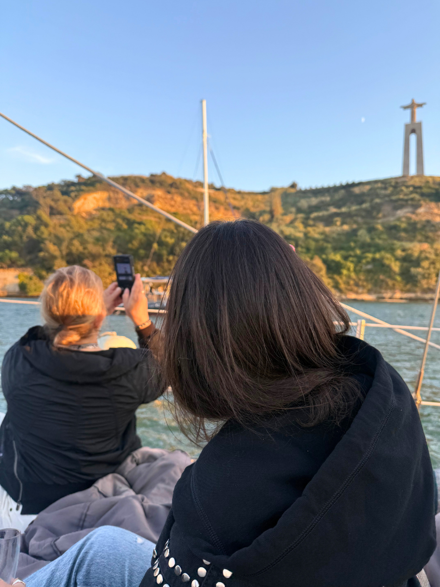 View from behind two women on a sailboat, one capturing a photo of the Cristo Rei statue on a hill near the Tagus River in Lisbon at sunset.