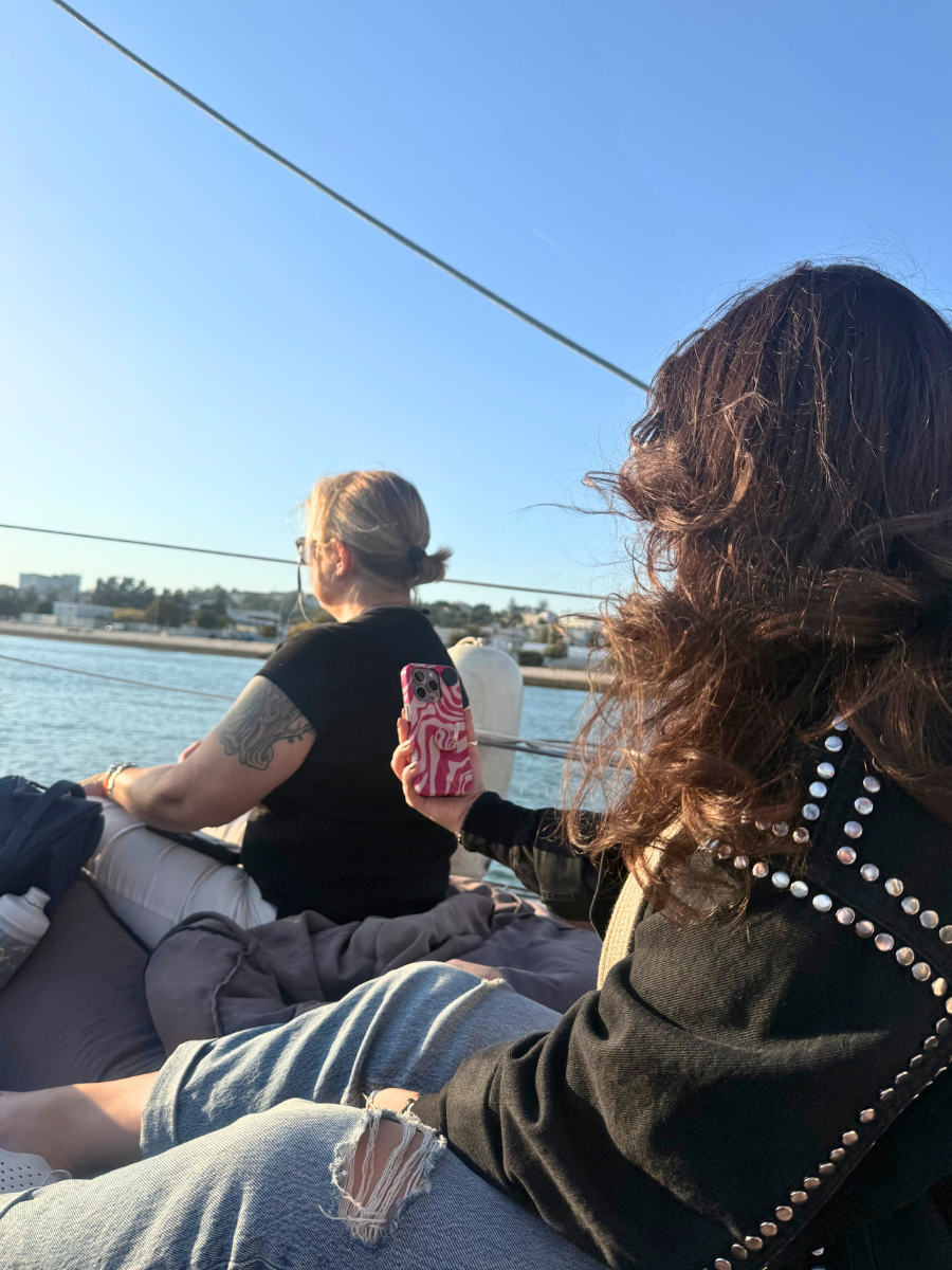 Two women sitting on a sailboat enjoying the ocean view; one is holding a patterned pink phone while looking toward the shoreline.
