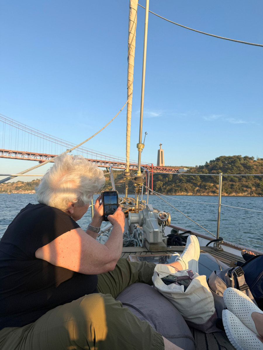 Woman taking a photo with her phone on a sailboat at golden hour, with the 25 de Abril Bridge and Cristo Rei statue in the background.