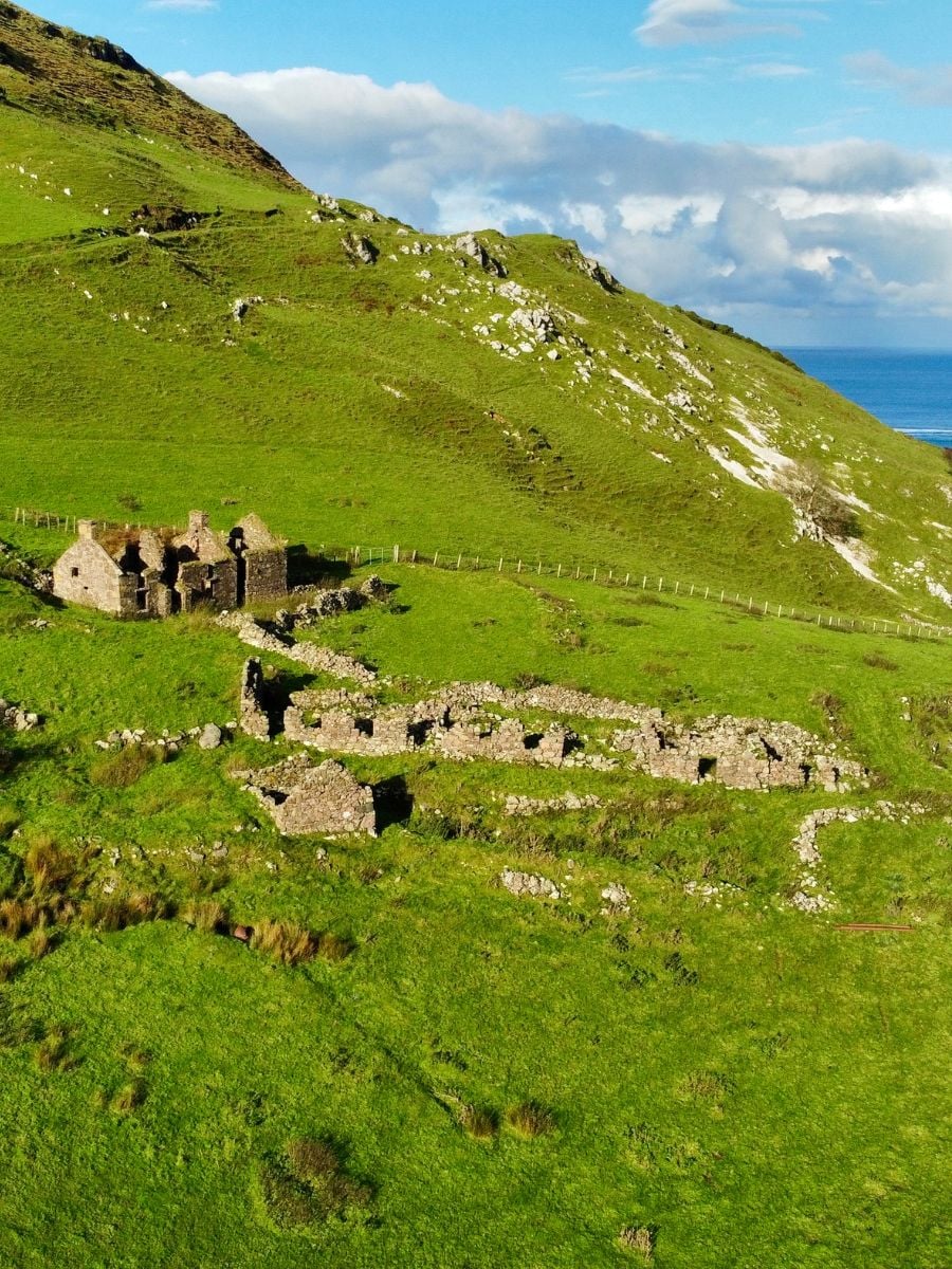 A scenic view of lush green hills along Ireland&rsquo;s coastline with the ruins of old stone cottages scattered across the landscape under a bright blue sky with scattered clouds.