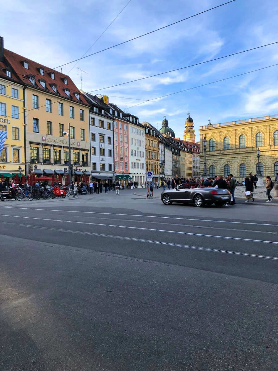 Street view in Munich with colorful historic buildings, people walking, and a busy city atmosphere under a blue sky.