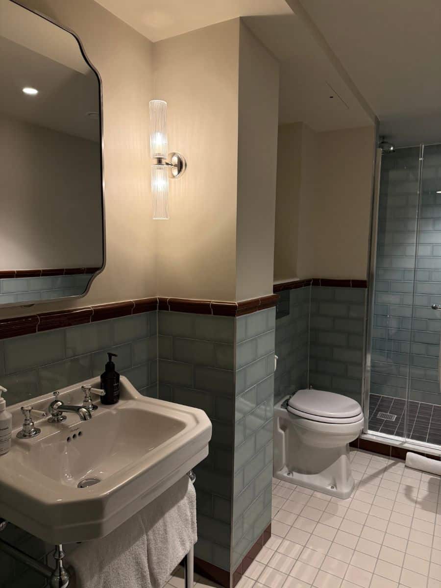 Bright hotel bathroom with a pedestal sink, tiled walls, glass shower, and classic fixtures.