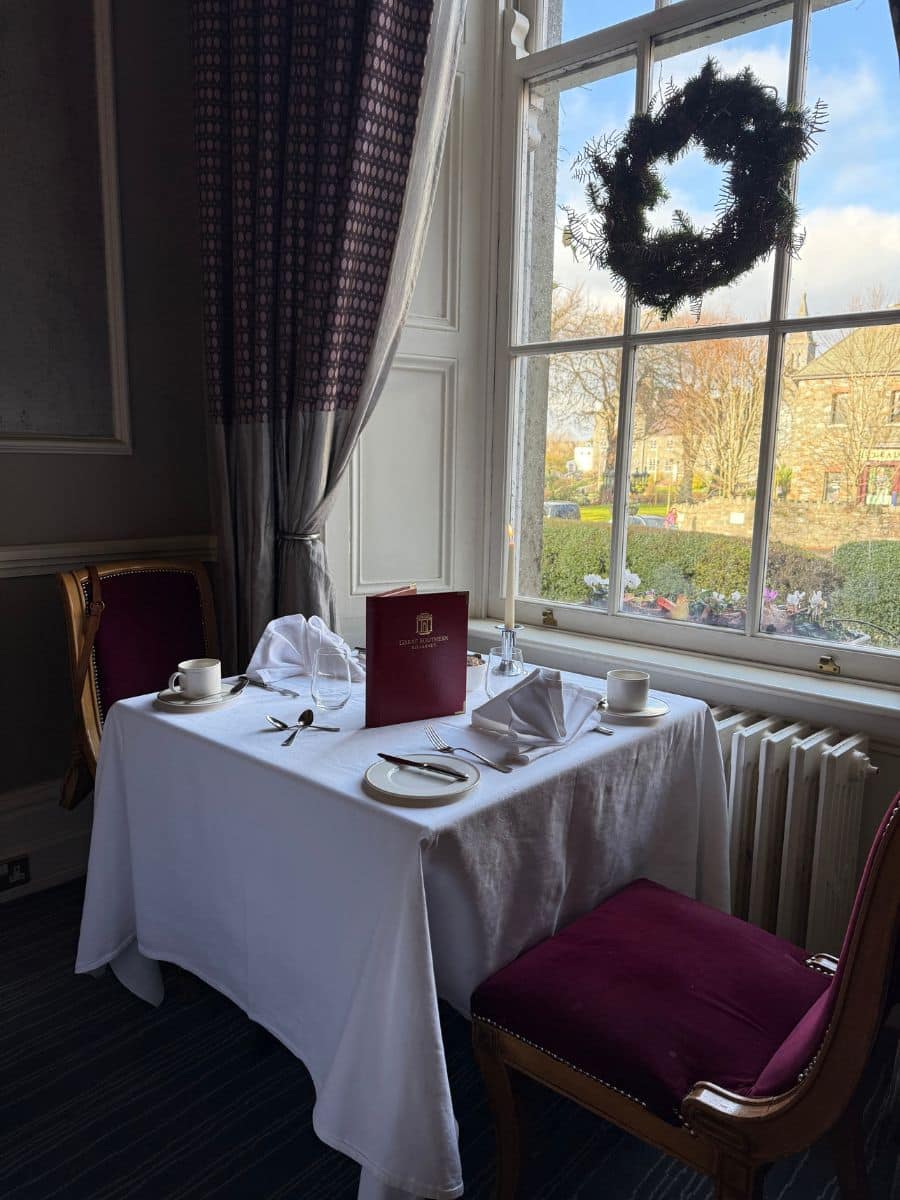 Elegant table set for afternoon tea beside a large window, decorated with a holiday wreath overlooking hotel gardens.