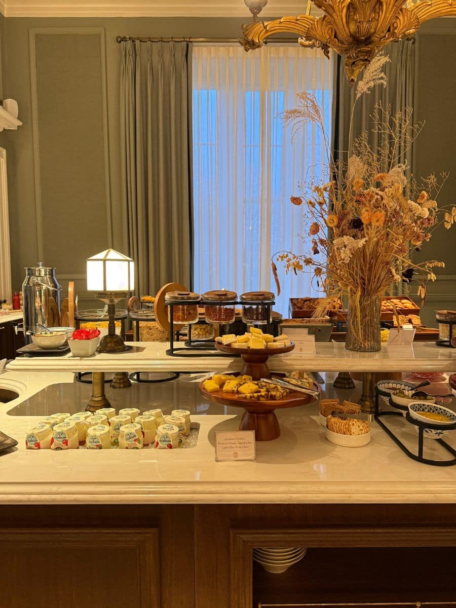 Hotel breakfast buffet featuring yogurt cups, pastries, cheeses, cereals, and dried flowers under warm lighting.