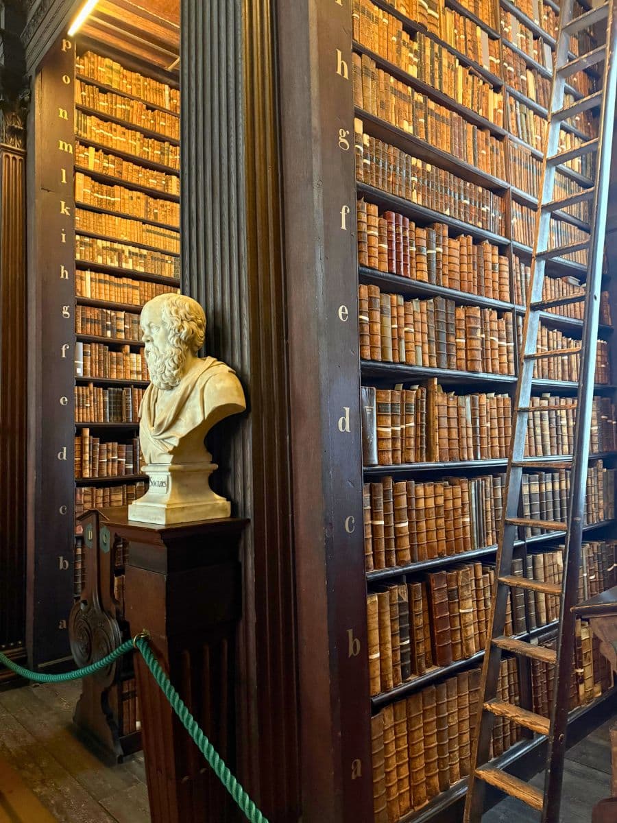 Interior of Trinity College’s Long Room with tall wooden bookshelves, a marble bust, and a classic library ladder.