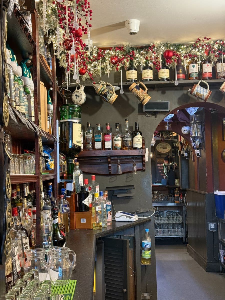 Festively decorated Irish pub bar lined with bottles of whiskey, hanging mugs, and Christmas garlands.