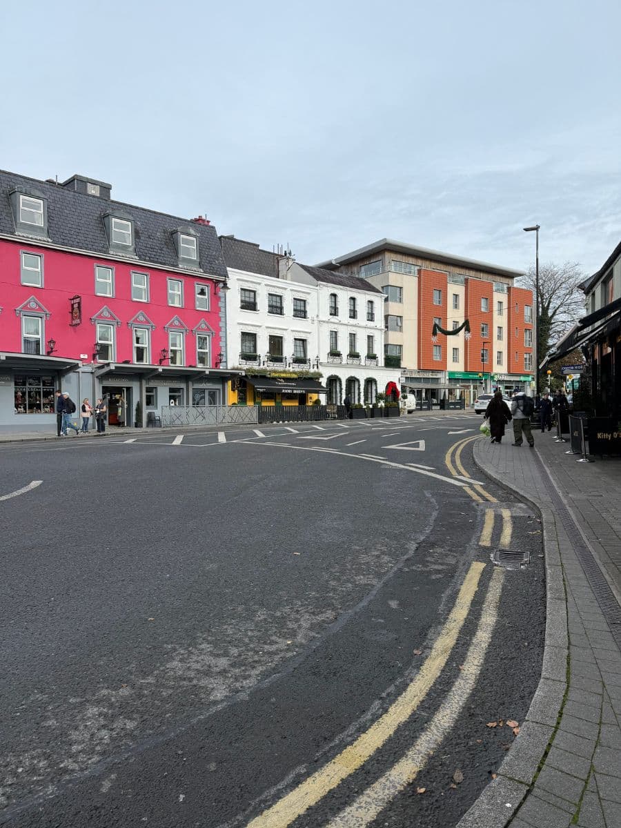 Street view of downtown Killarney featuring colorful shopfronts, including a bright pink building and traditional pubs.