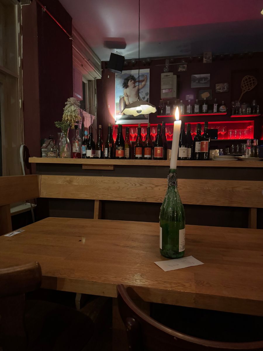 Cozy Berlin bar interior with a wooden table, a candle in a wine bottle, and a dimly lit bar lined with wine bottles and red backlighting.