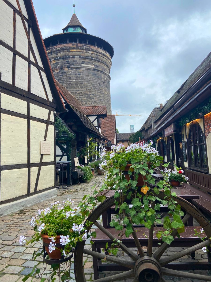 Charming cobblestone alley in Nuremberg’s Handwerkerhof lined with half-timbered houses, flower-filled planters, and the round medieval tower of the city wall in the background.