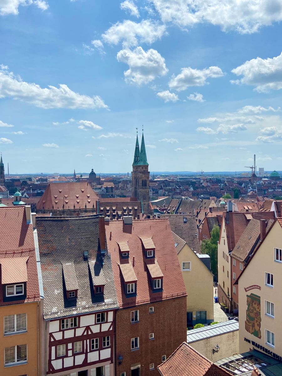 Panoramic view of Nuremberg’s old town with red-tiled rooftops, church spires, and historic buildings under a bright blue sky with scattered clouds.