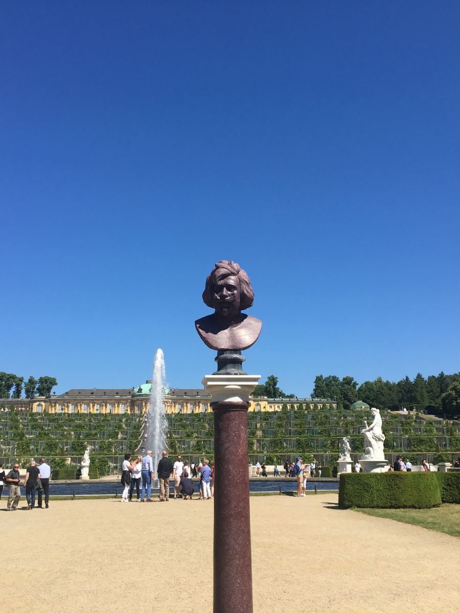 Bust statue on a tall pedestal with Sanssouci Palace and its cascading vineyard terraces in the background, under a clear blue sky in Potsdam, Germany.