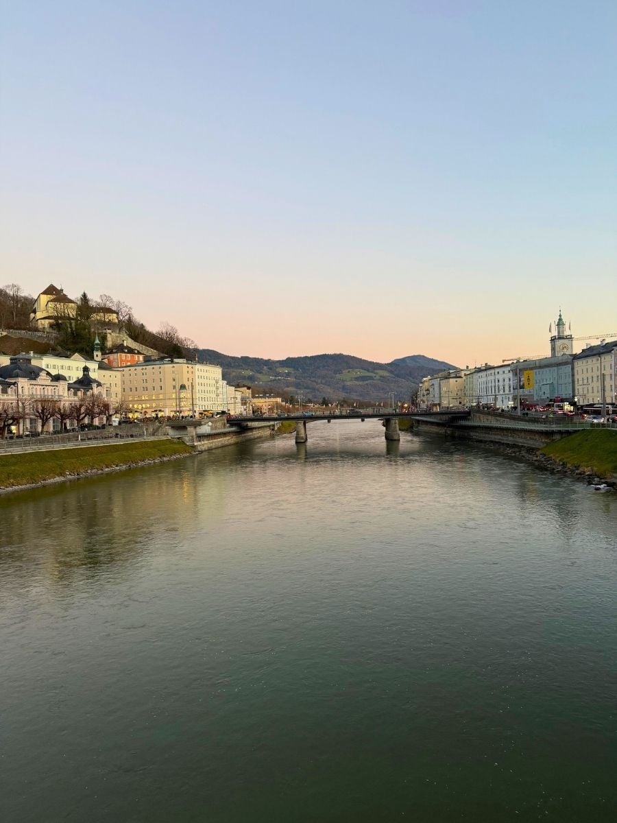 Riverside view of Salzburg with a bridge and pastel buildings at sunset.