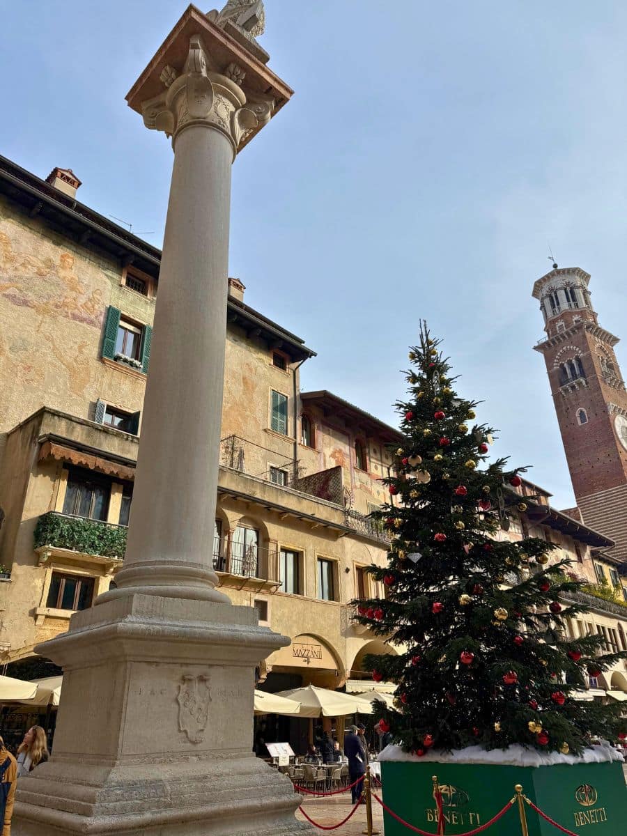 Tall column topped with the Lion of Saint Mark stands in Verona&rsquo;s Piazza delle Erbe beside a large Christmas tree decorated with red and gold baubles. The square is surrounded by old frescoed buildings and the Lamberti Tower is visible in the background under a clear blue sky.