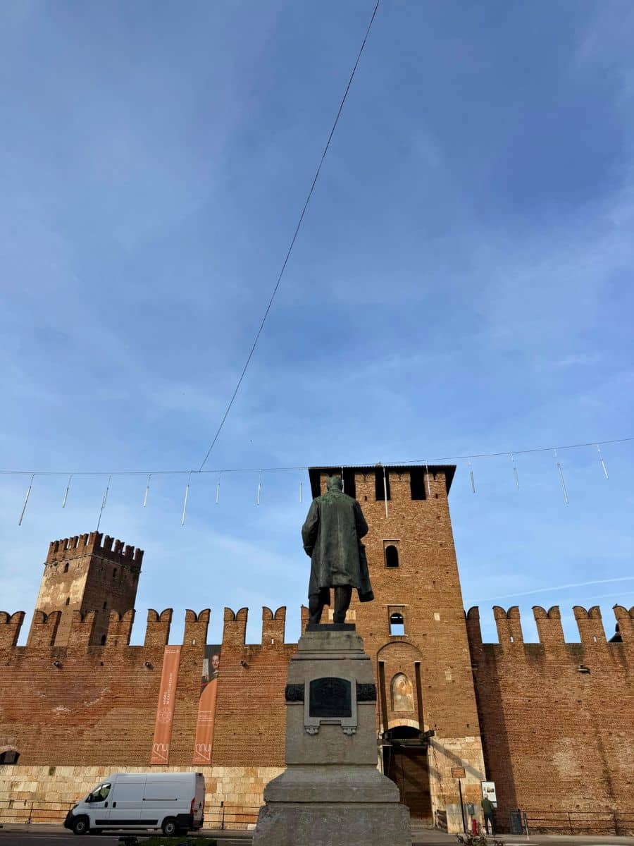 A statue facing the fortified walls and tower of Castelvecchio in Verona, Italy, under a blue sky with minimal clouds and light holiday decorations above.