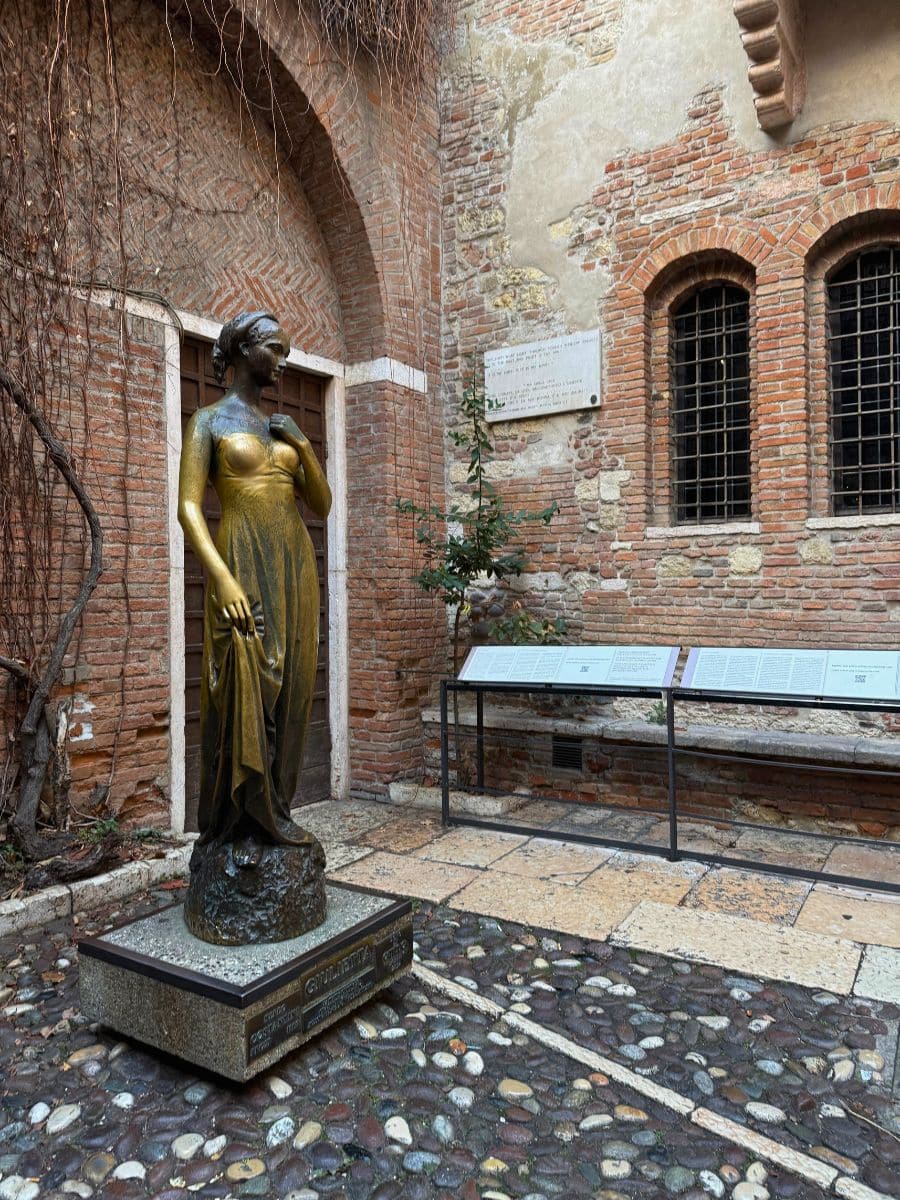 The bronze statue of Juliet in Verona&rsquo;s courtyard, standing in front of rustic red brick walls and surrounded by plaques and information boards.