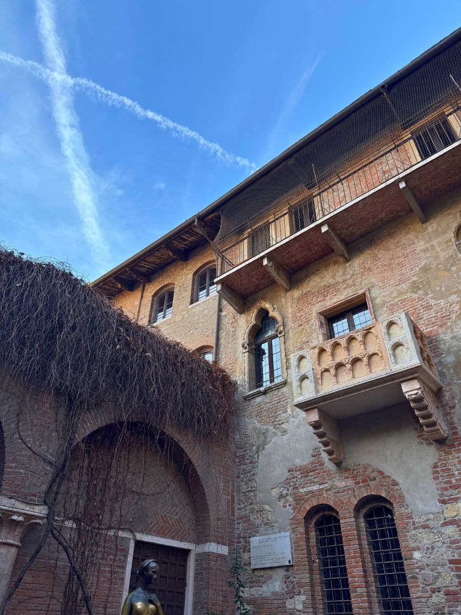 A view of Juliet&rsquo;s balcony in Verona, Italy, attached to a rustic stone building with wooden windows and climbing vines, under a bright blue sky.