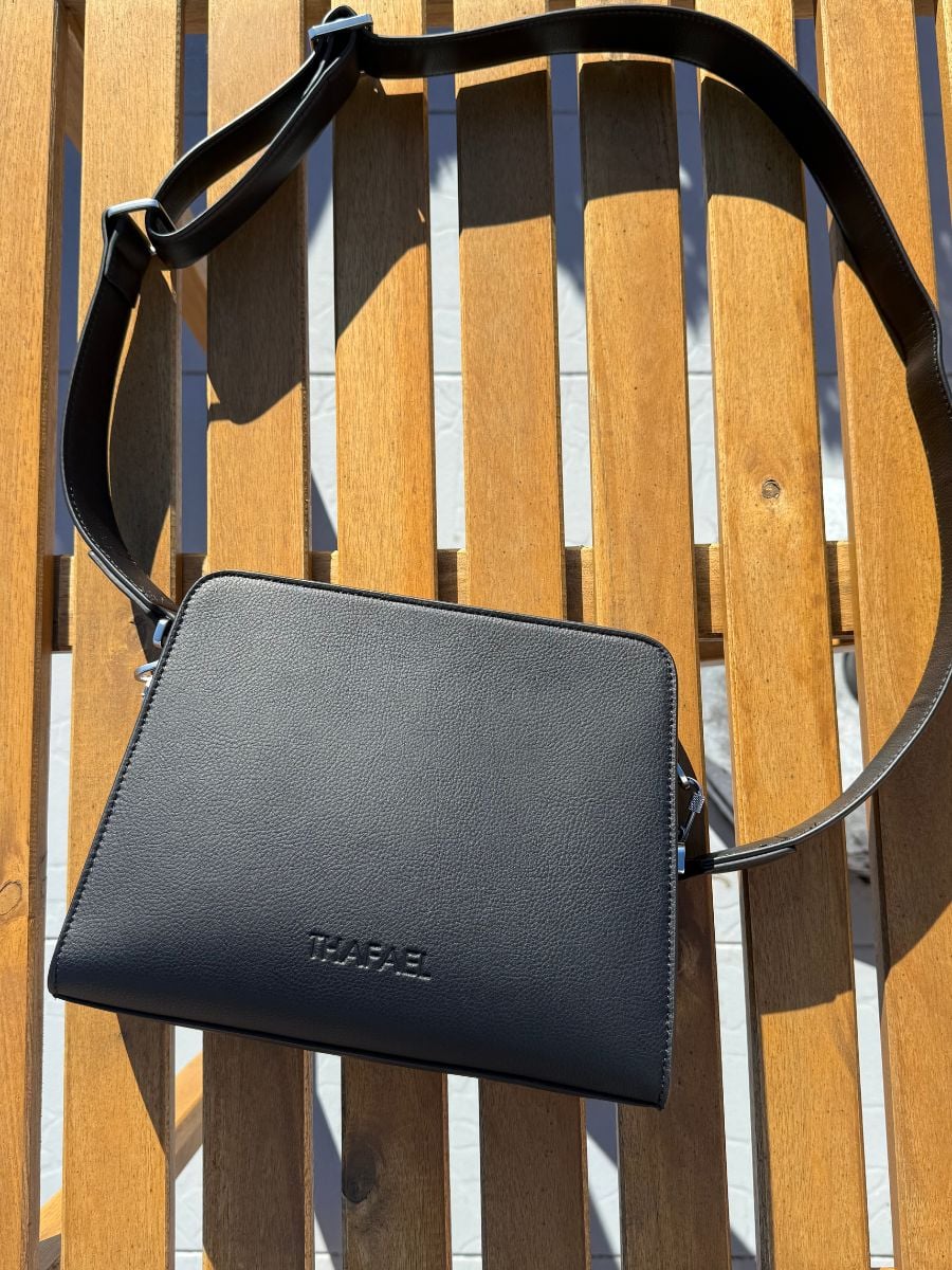 Black leather crossbody bag with adjustable strap resting on a wooden outdoor chair in sunlight.