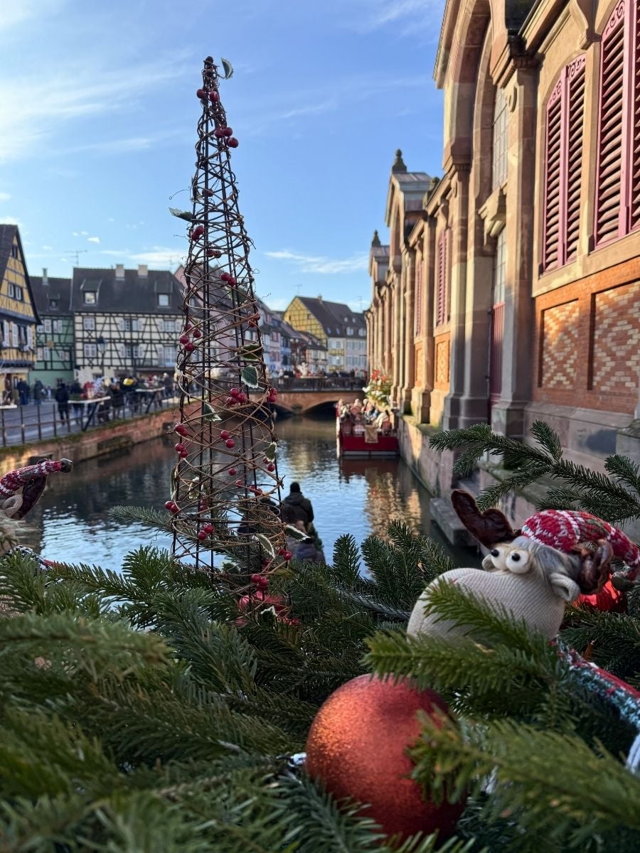 Festive canal view in Colmar with Christmas decorations, half-timbered houses, and holiday ornaments in foreground