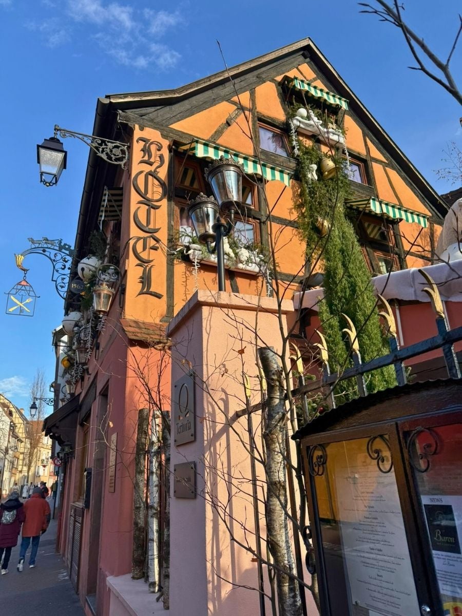 Colorful half-timbered hotel in Strasbourg with warm orange facade, green striped awnings, decorative lanterns, and winter greenery under a clear blue sky.
