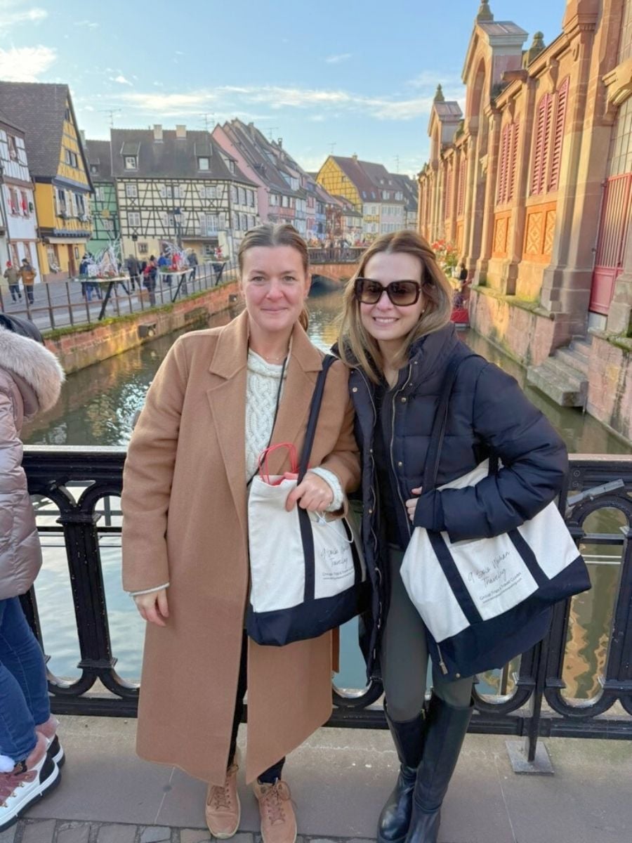 Two women smiling on a bridge in Strasbourg with canal and traditional Alsatian half-timbered houses in the background during golden hour.
