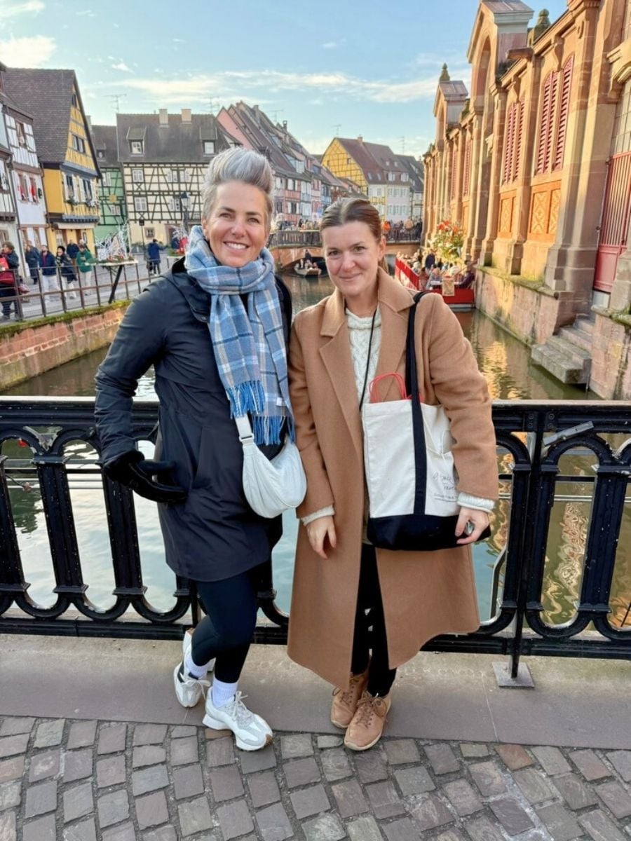 Two friends posing on a Strasbourg bridge with canal reflections and pastel half-timbered buildings behind them on a crisp winter afternoon.