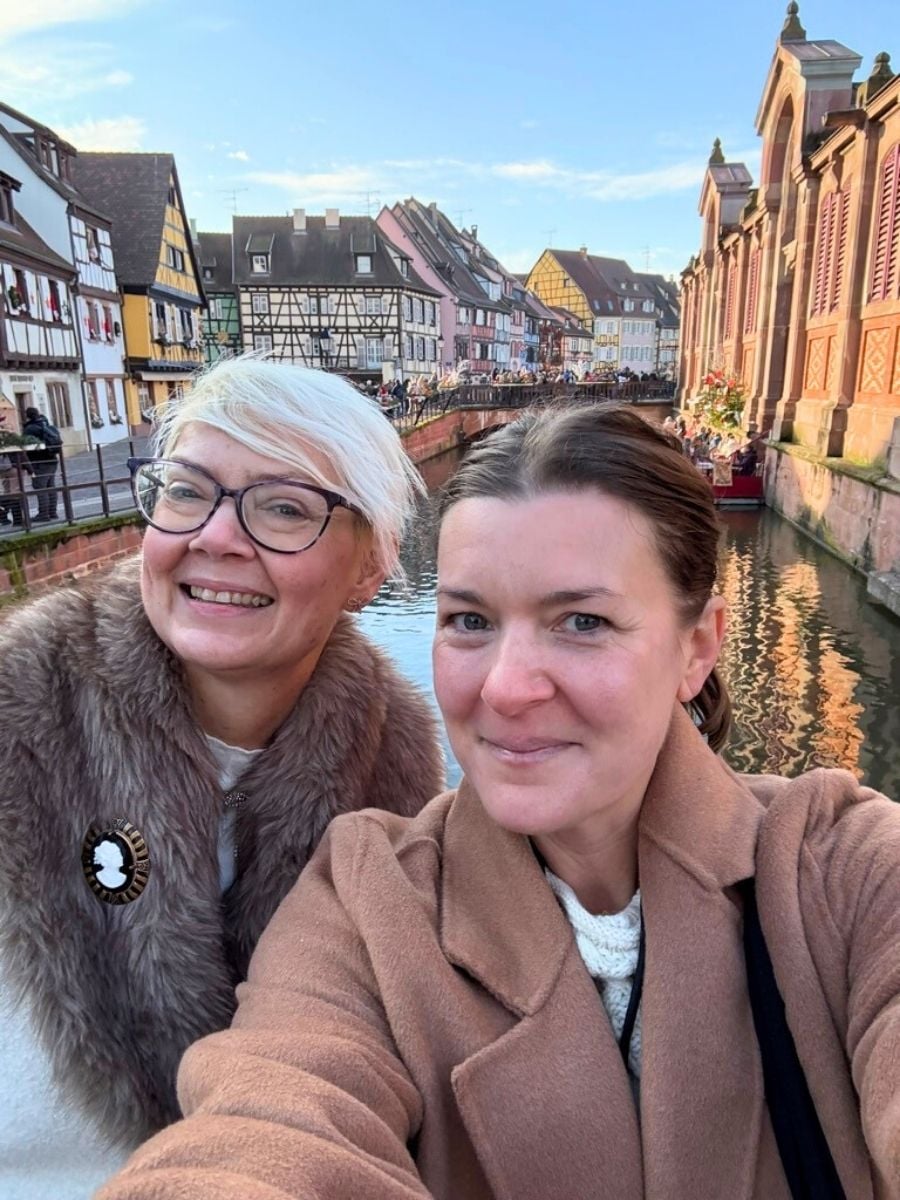 Close-up selfie of two women smiling on a Strasbourg bridge with canal and historic timber-framed buildings in the background.