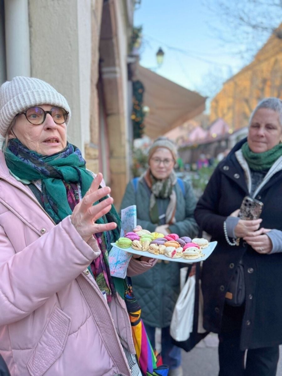 Local guide holding a tray of colorful macarons while speaking to a small group during a Strasbourg food tour in winter.