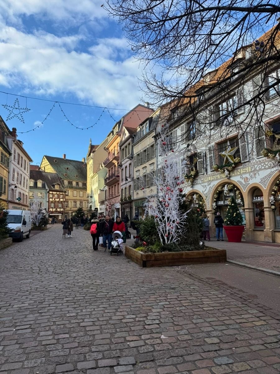 Cobblestone street in Colmar, France with colorful half-timbered houses, Christmas decorations, and people walking under a blue sky.