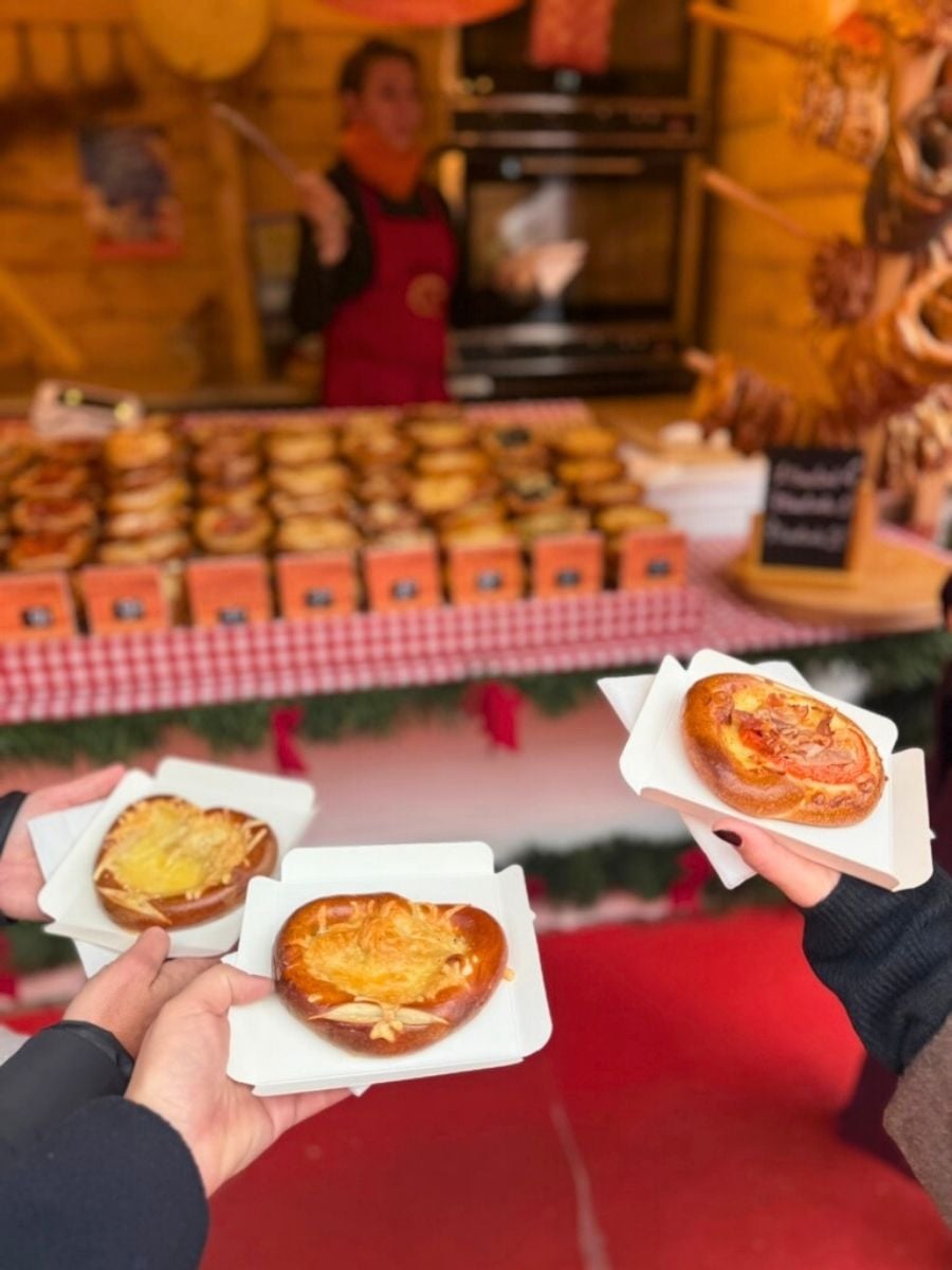 Three hands holding warm cheese-topped pretzels at a Christmas market stall with baked goods displayed in the background.