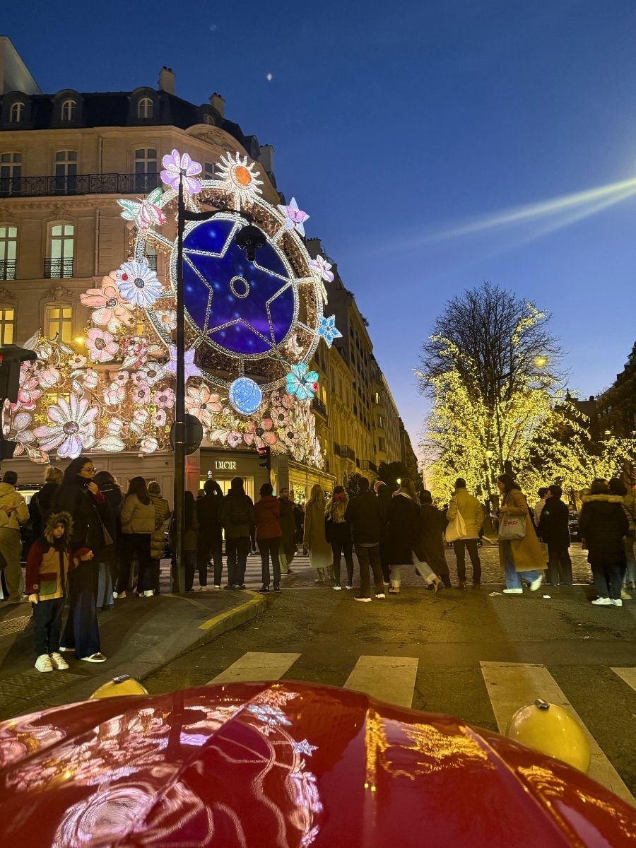 Crowd gathered at a Paris street corner at night with colorful Christmas light decorations and illuminated trees.
