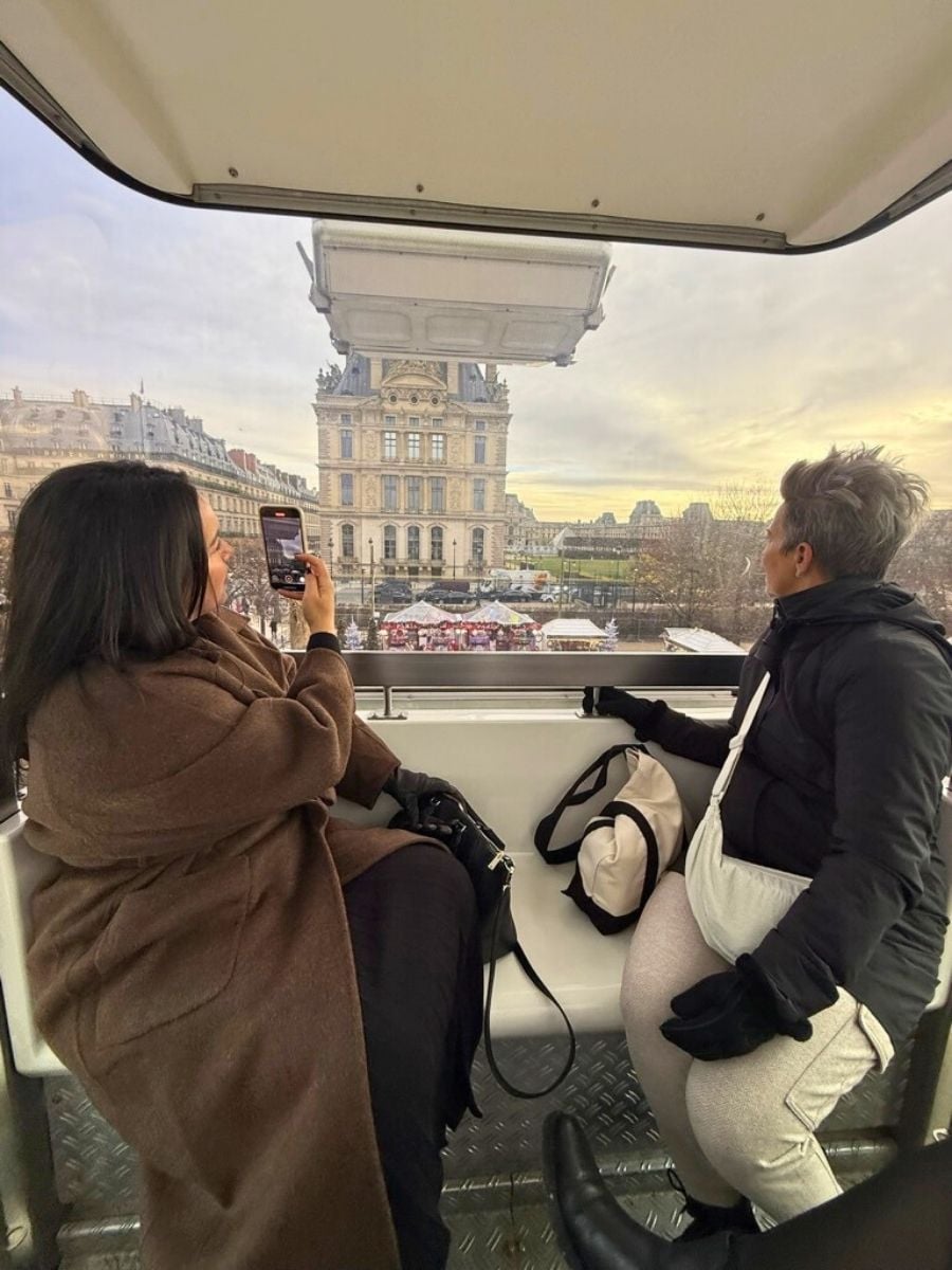 Two women sitting inside a Ferris wheel cabin overlooking the Louvre Museum and Christmas market in Paris at sunset.