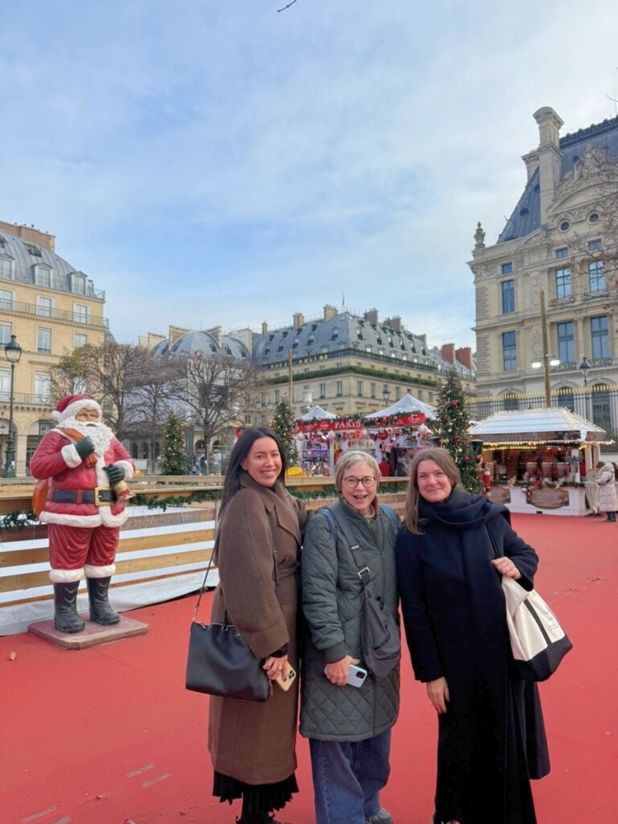 Three women smiling at a Christmas market in Paris with festive stalls and Santa statue in the background