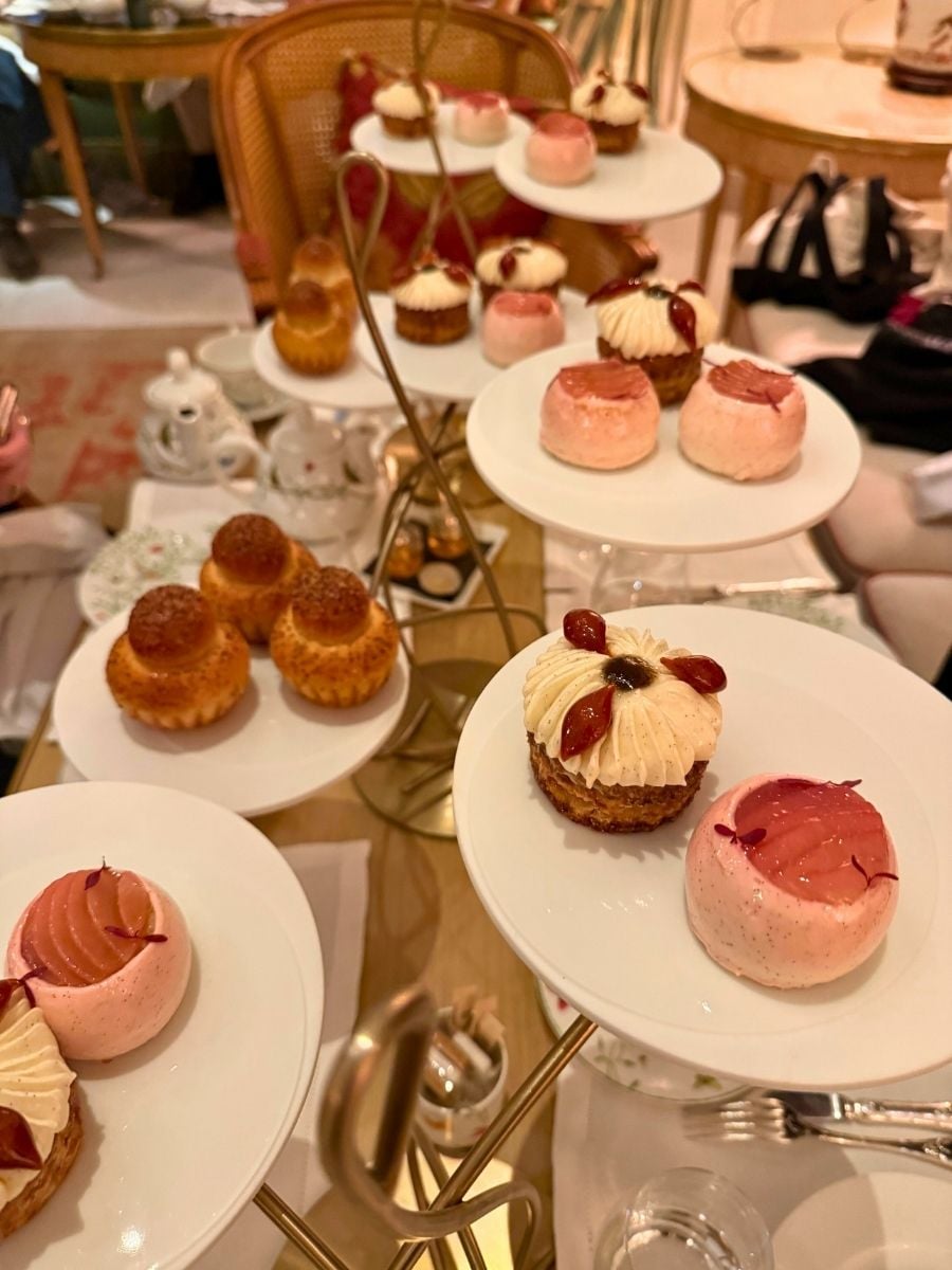Tiered afternoon tea stand with pink pastries, cream-topped mini cakes, and golden brioche in an elegant Paris caf&eacute; setting.