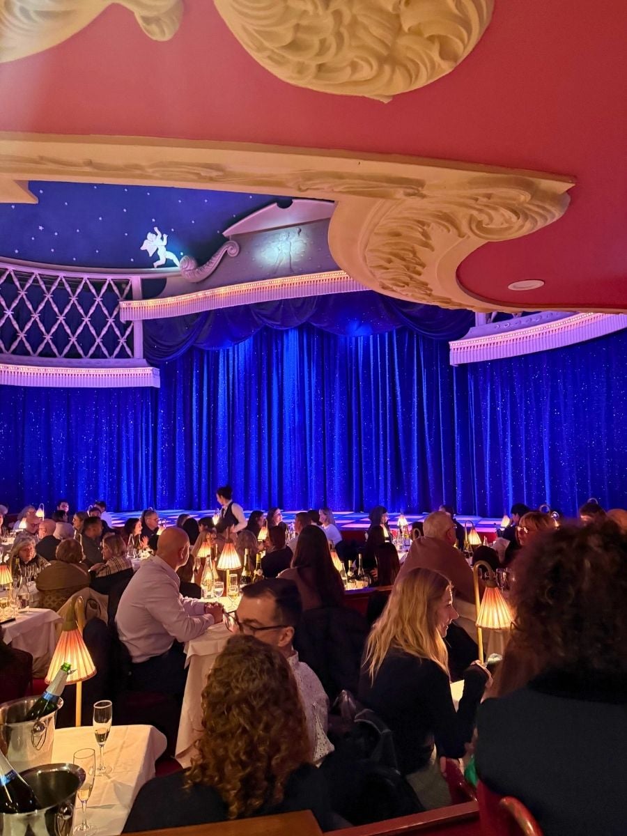 Elegant Paris cabaret-style dinner show venue with blue velvet stage curtains and guests seated at candlelit tables.